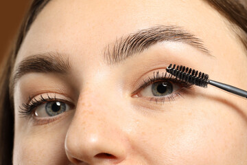 Naklejka premium Woman applying mascara onto her eyelashes on brown background, closeup