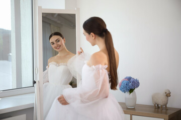 Dreams come true. Woman wearing beautiful wedding dress looking at herself in mirror indoors