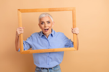 Elderly woman holding a wooden frame, showcasing surprise expression, wearing blue striped shirt,...