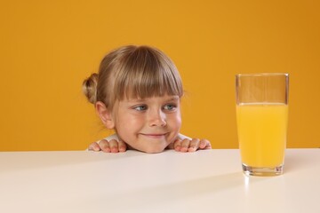 Cute little girl looking at glass of juice while peeking over table against orange background