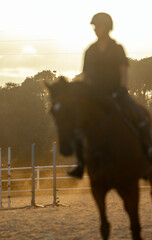 Close-up of a blurred backlit silhouette of a woman riding a horse at sunset, with the glow of the setting sun in the background. © sirbouman