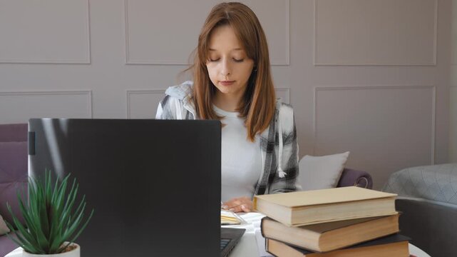Young female student diligently taking notes from laptop screen, writing in notebook while preparing for exams and studying at home with focused concentration