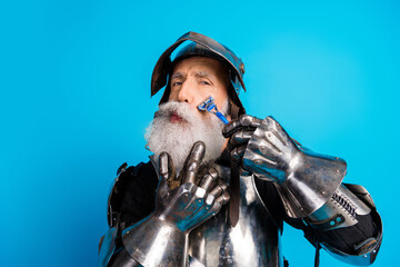Fototapeta premium Elder knight in medieval armor whimsically shaving against blue background, showcasing humor and historical charm.