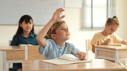 Fototapeta premium Caucasian smart boy raising hand for asking and answering teacher surrounded by diverse children studying at classroom while teacher walking pass at elementary classroom. Education concept. Pedagogy.