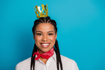Young woman with a charming style and braided hair wearing a crown and scarf smiling against a blue...