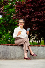 Obraz premium Mature businesswoman enjoying coffee outdoors, holding smartphone in relaxed yet professional urban setting on a warm day