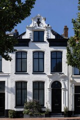 Obraz premium White brick house with large windows and unique architectural features located on a sunny street surrounded by trees, ideal for a peaceful neighborhood stroll