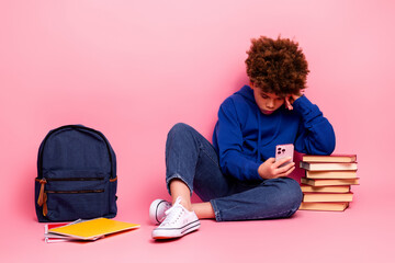 Young boy sitting on the floor using smartphone beside stack of book and backpack over pink background