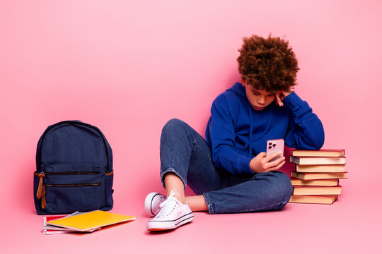 Young boy sitting on the floor using smartphone beside stack of book and backpack over pink background - Powered by Adobe