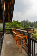 A stylish outdoor cafe balcony with a row of wooden bar stools and a long counter overlooking a lush tropical landscape and the open sky.
