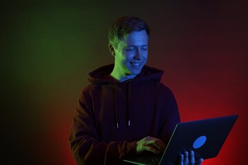 Smiling man wearing hoodie using laptop in neon lights against black background