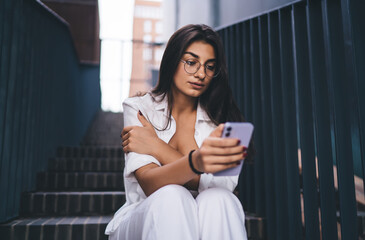 Thoughtful brunette woman sitting with phone, absorbed in content, showing connection to digital...