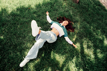 Young woman lying on grass outdoors in trendy casual outfit during spring, expressing joy and...
