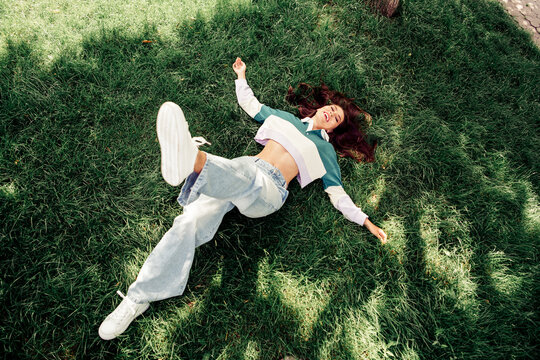 Young woman lying on grass outdoors in trendy casual outfit during spring, expressing joy and relaxation in natural light
