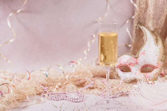 Masquerade ball holiday background. Glass of white champagne, Venetian carnival mask and feathers on light pink background