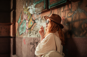 Stylish red-haired woman smoking in urban street with graffiti