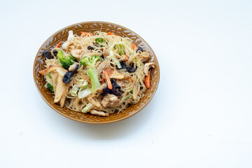 A bowl of stir-fried rice vermicelli with chicken, broccoli, carrots, and mushrooms. Traditional Asian noodle dish, colorful and healthy meal served on white background.