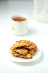 A small stack of fried banana fritters on a white plate, with a blurry cup of tea and a teapot in the background.