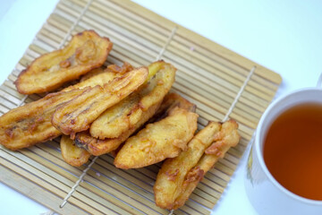 A small pile of fried banana fritters sits on a bamboo mat next to a white cup of hot tea.