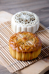 A golden baked mooncake sits in front of a snow skin mooncake on a bamboo mat, highlighting two traditional Mid-Autumn pastries with intricate floral designs.