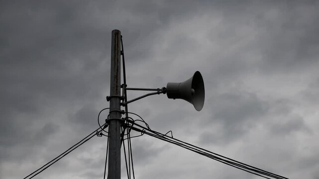 single public address speaker mounted high upon vertical pole with connecting cables visible against overcast sky filled with clouds
