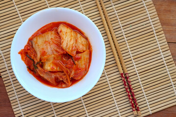 A white bowl filled with spicy red kimchi sits on a woven bamboo mat next to a pair of chopsticks on a wooden surface.