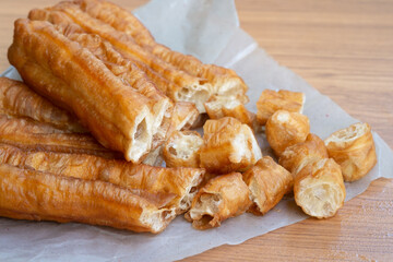 Long golden fried dough sticks lying on paper with smaller cut pieces scattered nearby, showing their crisp shell and soft airy inside on a wooden table.