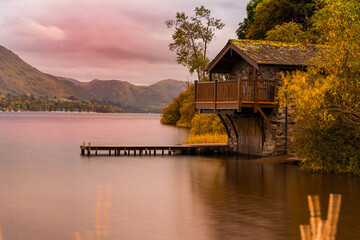 Obraz premium Stone boathouse at Ullswater in the Lake District with golden autumn trees and still lake water under a pastel evening sky.