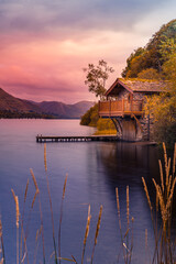 Obraz premium Soft pink and purple sunset sky above a stone boathouse on Ullswater lake, framed by autumn grasses and trees.