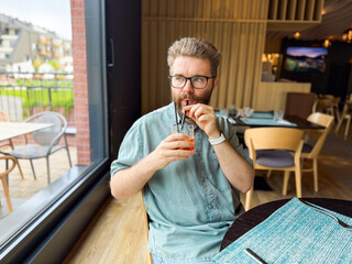 Man drinking cocktail with straw while sitting near restaurant window. Refreshment, leisure, and hospitality lifestyle captured in casual dining environment.