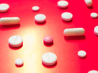 On a bright red background, white and one pink tablet, as well as capsules, are scattered at...