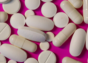 A variety of white capsules and tablets on a vibrant fuchsia background create a dynamic and contrasting composition. The photo illustrates themes of health, medicine, and pharmaceuticals, highlightin