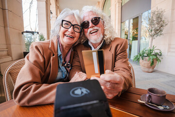 Cheerful senior couple joyfully capturing a selfie at a cafe, proudly displaying their credit card as they cherish their time together, showcasing love and connection during a delightful outing filled