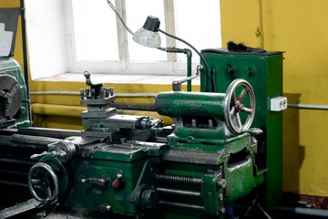 A large, vintage green radial arm drill press dominates a small workshop. Metalworking tools and drill bits rest on a nearby table, near a gray wall and a yellow wall