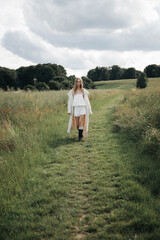 oung woman walking outdoors in a meadow wearing a long knitted cardigan, white lace outfit, and black boots, enjoying a peaceful summer day 