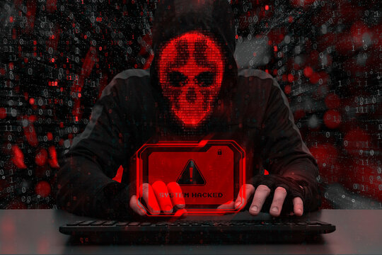 Masked hacker behind a computer with a red warning System hacked. Concept of cybersecurity threat, attacks on data and online systems.