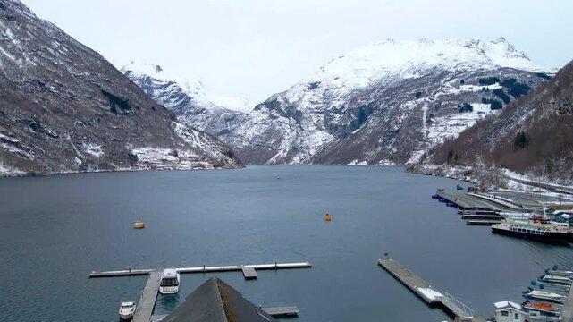 Panoramic view of the majestic Geirangerfjord and the charming village of Geiranger in winter, surrounded by snow capped mountains and calm, deep blue waters.