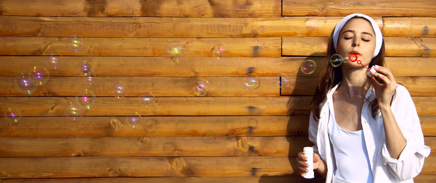 front view shot of young female model blowing soap bubbles on wooden board wall of house background. Playful girl woman blow party bubbles  Lady celebrates a special occasion or birthday, Lightness