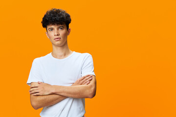 serious young man standing with crossed arms against bright orange background wearing casual white...