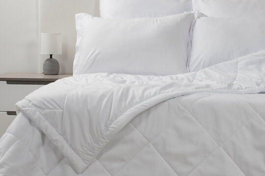 A bed with white pillows and a blanket, next to a bedside table with a lamp, daylight