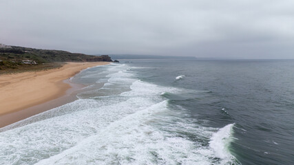 White waves gently crash on a sandy beach in Nazare, Portugal, under an overcast sky. The serene...