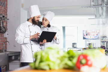 In a luxurious equipped kitchen, team of chefs in uniform using a laptop to find new cooking ideas or tips. Culinary professionals doing food prep with raw materials ingredients, chef life.