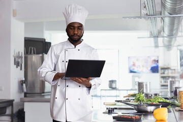 African american chef researching new cooking tips in restaurant kitchen uses a laptop while...