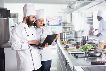 Head chefs in a white uniform researching new gourmet recipes on a laptop while overseeing food...