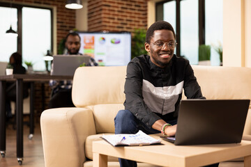 African american marketing specialist seated with laptop and clipboard, reviewing startup company analysis. Black male manager on cozy sofa, using his personal computer in brick wall office.