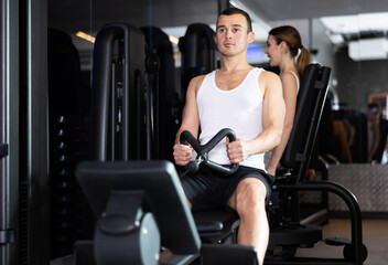 Fototapeta premium Male gym visitor exercises on simulator in order to strengthen muscles of arms. He pulls handle of simulator and drains muscles of his arms.