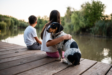 Children hugging dog and relaxing by lake on pier at sunset
