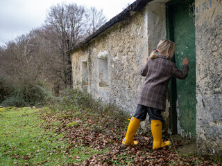 Blonde girl wearing yellow boots knocking on the weathered door of an old, abandoned house nestled...