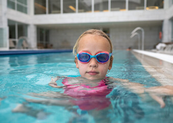 Portrait of a young girl enjoying a swim in an indoor pool, wearing colorful goggles and surrounded...