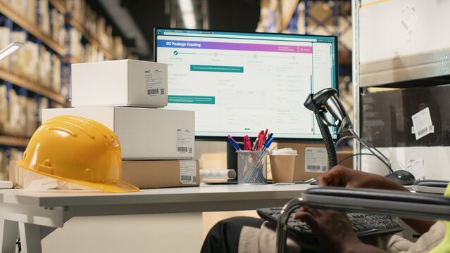 African american male staff with disability uses airway bill numbers for tracking info on software, monitoring the parcel status on computer in logistics hub. Shipment operations. Camera B.